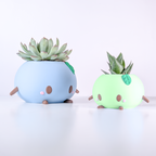May include: Two round planters shaped like creatures, one light blue and one light green. Each features small brown limbs, brown oval eyes, and pink circular cheeks. The blue planter holds a large succulent, while the green planter holds a spiky succulent.