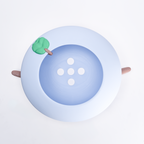A top-down view of a light blue circular button with a slightly darker blue center, featuring four white circular buttonholes and a small green and brown leaf detail on the upper left edge. Two small brown sticks extend from the sides.