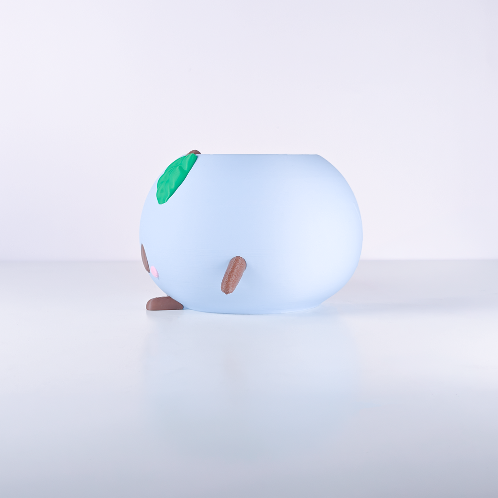 A light blue, rounded, cartoon-like decorative object with small brown legs and a green leaf detail on top, photographed on a clean white studio surface.