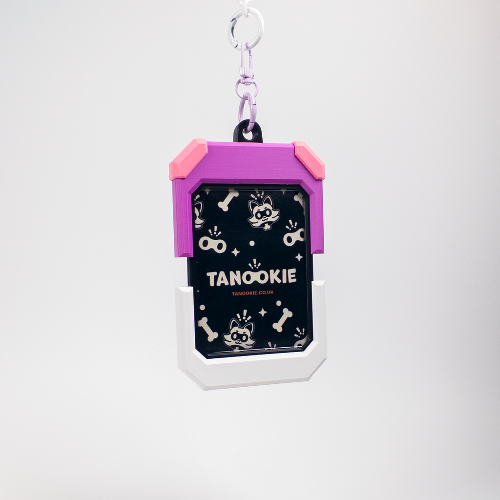A Tanookie brand keychain or ID holder with a purple and pink frame, a white lower border, and a black center panel decorated with white dog-themed graphics and the text 'TANOOKIE'. It is attached to a silver metal ring and clasp, shown against a plain light gray background.
