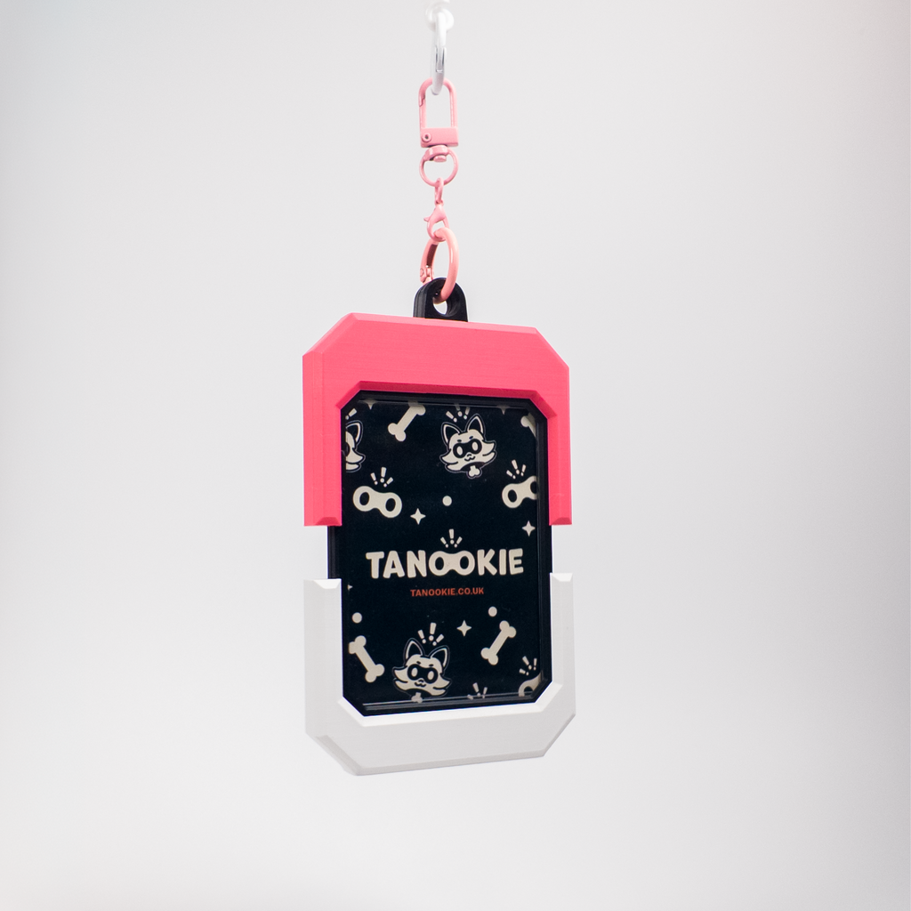 A pet ID tag keychain with a pink top section, a black middle section featuring a raccoon pattern and the text 'TANOOKIE', and a white bottom section, hanging from a pink clip against a light gray background.