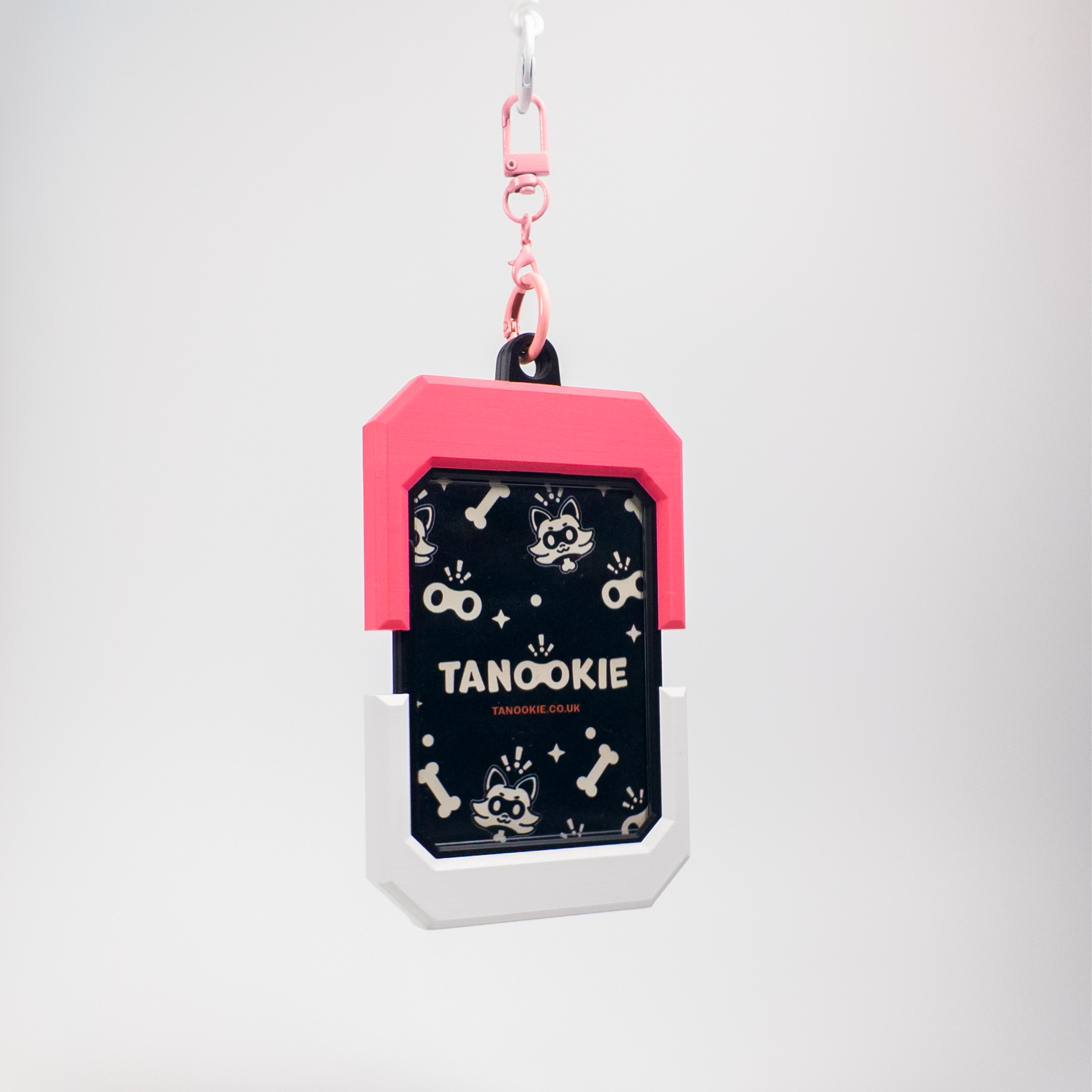 A pet ID tag keychain with a pink top section, a black middle section featuring a raccoon pattern and the text 'TANOOKIE', and a white bottom section, hanging from a pink clip against a light gray background.