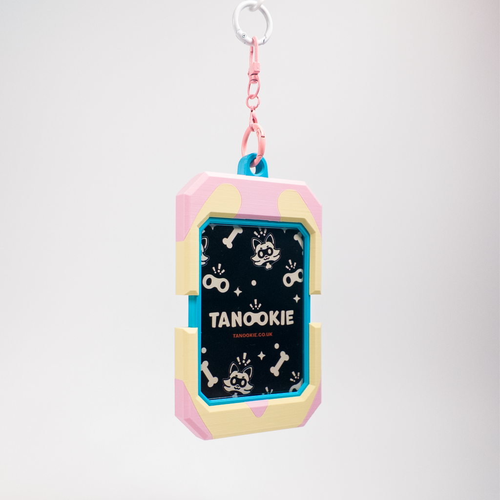 A cute, pastel-colored dog keychain or bag charm featuring a pink and cream shell-like frame with a black center printed with white dog illustrations and the text 'TANOOKIE'. It has a pink chain and a silver metal keyring at the top, set against a clean, light gray gradient background.