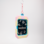 A cute, pastel-colored dog keychain or bag charm featuring a pink and cream shell-like frame with a black center printed with white dog illustrations and the text 'TANOOKIE'. It has a pink chain and a silver metal keyring at the top, set against a clean, light gray gradient background.
