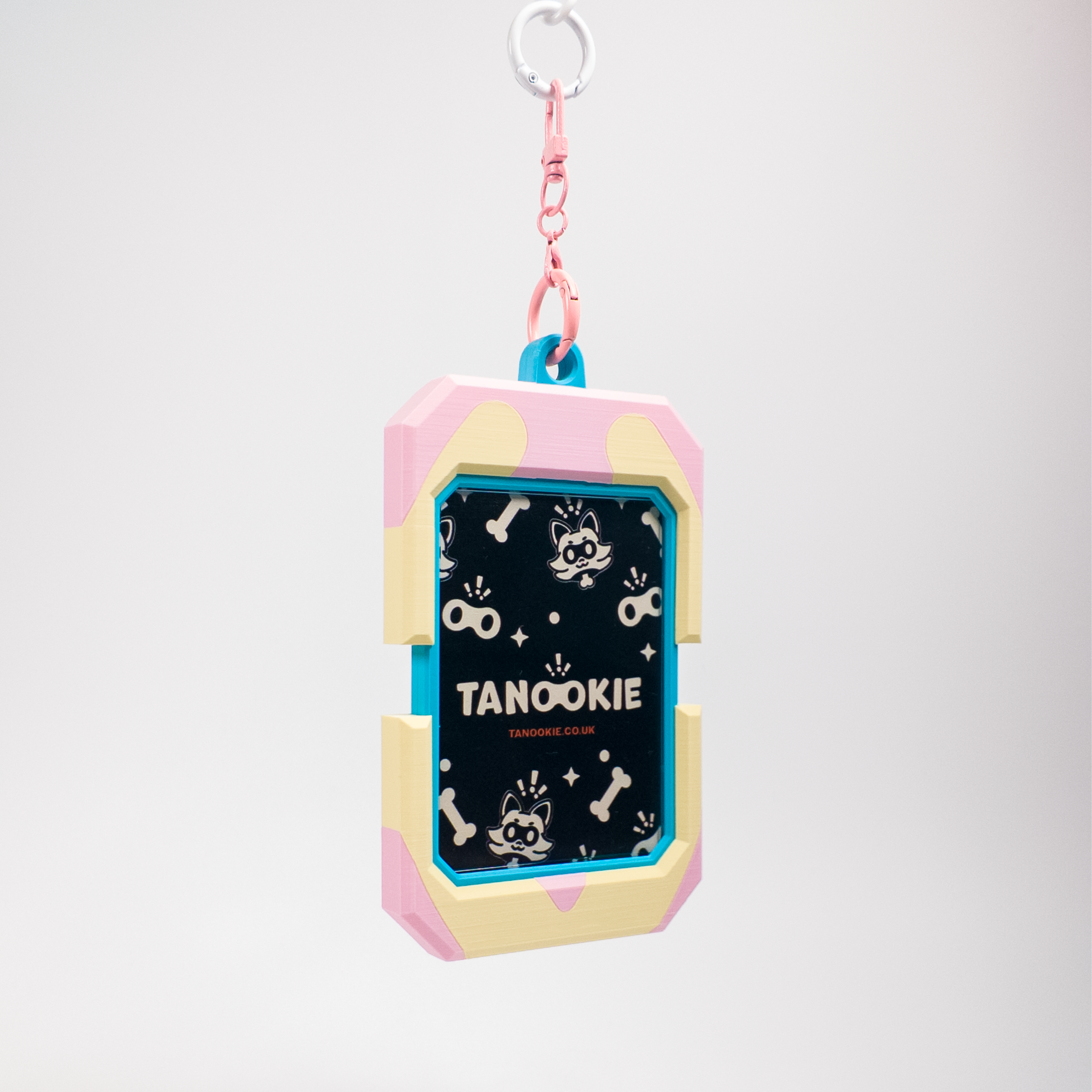 A cute, pastel-colored dog keychain or bag charm featuring a pink and cream shell-like frame with a black center printed with white dog illustrations and the text 'TANOOKIE'. It has a pink chain and a silver metal keyring at the top, set against a clean, light gray gradient background.