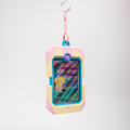 A pastel-colored card holder or game card case with a pink and yellow frame, turquoise trim, and a holographic card insert, hanging from a silver carabiner on a light gray background.