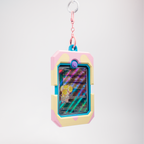 A pastel-colored card holder or game card case with a pink and yellow frame, turquoise trim, and a holographic card insert, hanging from a silver carabiner on a light gray background.