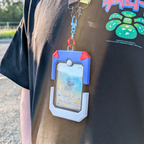 A close-up lifestyle shot of a person wearing a black t-shirt and holding a blue and white holographic Pokémon card holder with red accents. The holder is attached to a lanyard with a silver clasp.