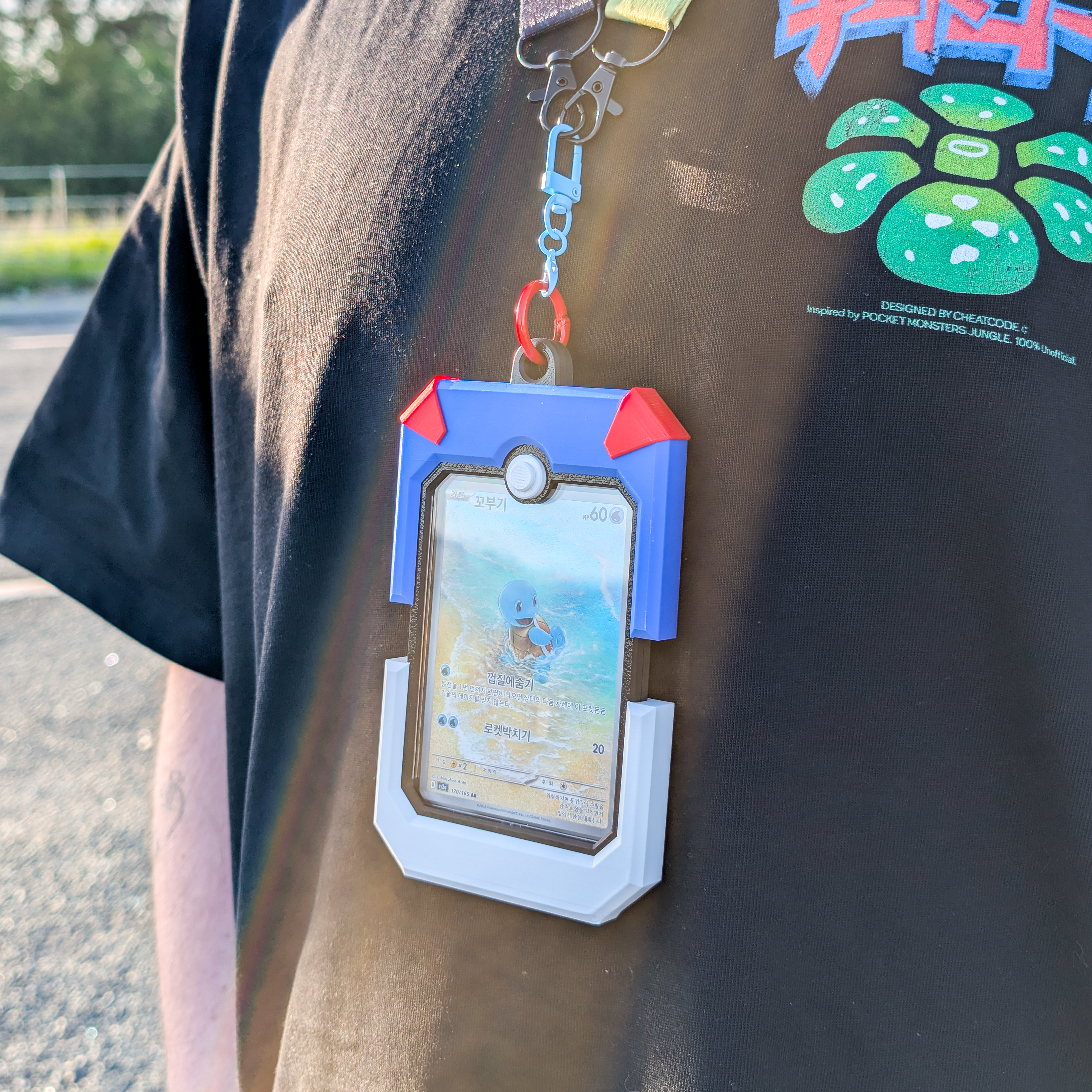 A close-up lifestyle shot of a person wearing a black t-shirt and holding a blue and white holographic Pokémon card holder with red accents. The holder is attached to a lanyard with a silver clasp.