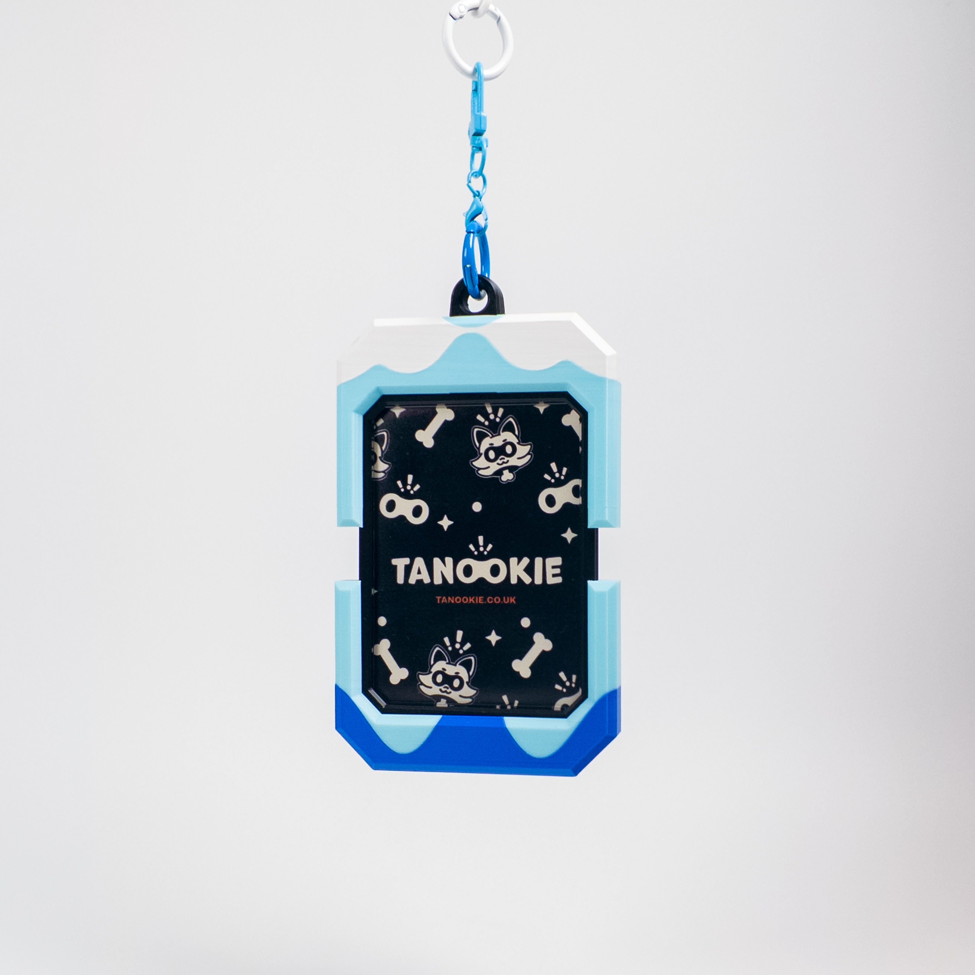 A dog ID tag keychain featuring a black center with white dog-themed illustrations and the text 'TANOOKIE' and 'TANOOKIES', surrounded by light blue and royal blue borders, suspended from a blue chain on a light gray background.