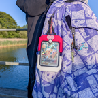 A close-up lifestyle shot of a purple and white patterned backpack with a red and white protective card holder containing a Pokémon TCG card, hanging outdoors next to a river.