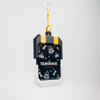 A rectangular keychain or bag charm with a dark navy front panel printed with white cartoon skulls, bones, and the word 'TANOOKIE' in white, attached to a yellow clasp and silver key ring on a light gray background.