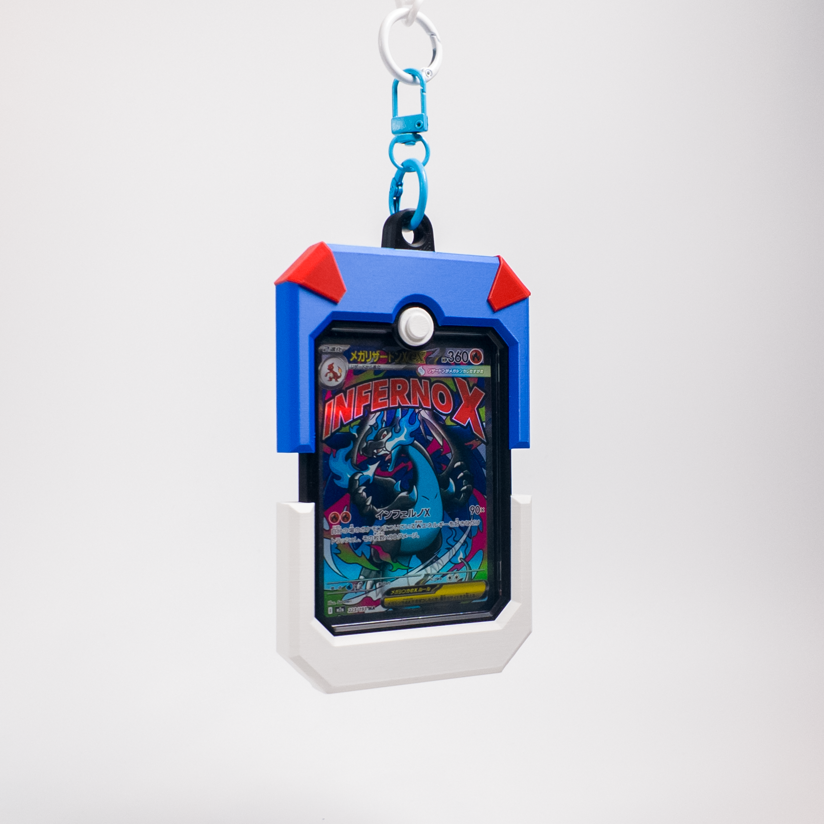 A blue and white card holder keychain case with red corner accents, holding a colorful 'Inferno X' trading card, suspended from a blue and silver carabiner on a light gray background.