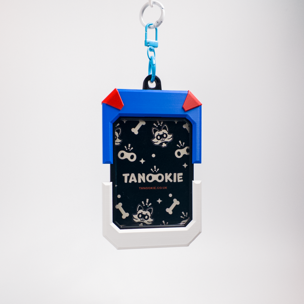 A Tanookie dog ID tag keychain featuring a blue and white rugged frame with red corner accents, a black patterned center with dog illustrations, and a blue carabiner clip at the top, shown on a light gray background.