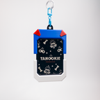 A Tanookie dog ID tag keychain featuring a blue and white rugged frame with red corner accents, a black patterned center with dog illustrations, and a blue carabiner clip at the top, shown on a light gray background.