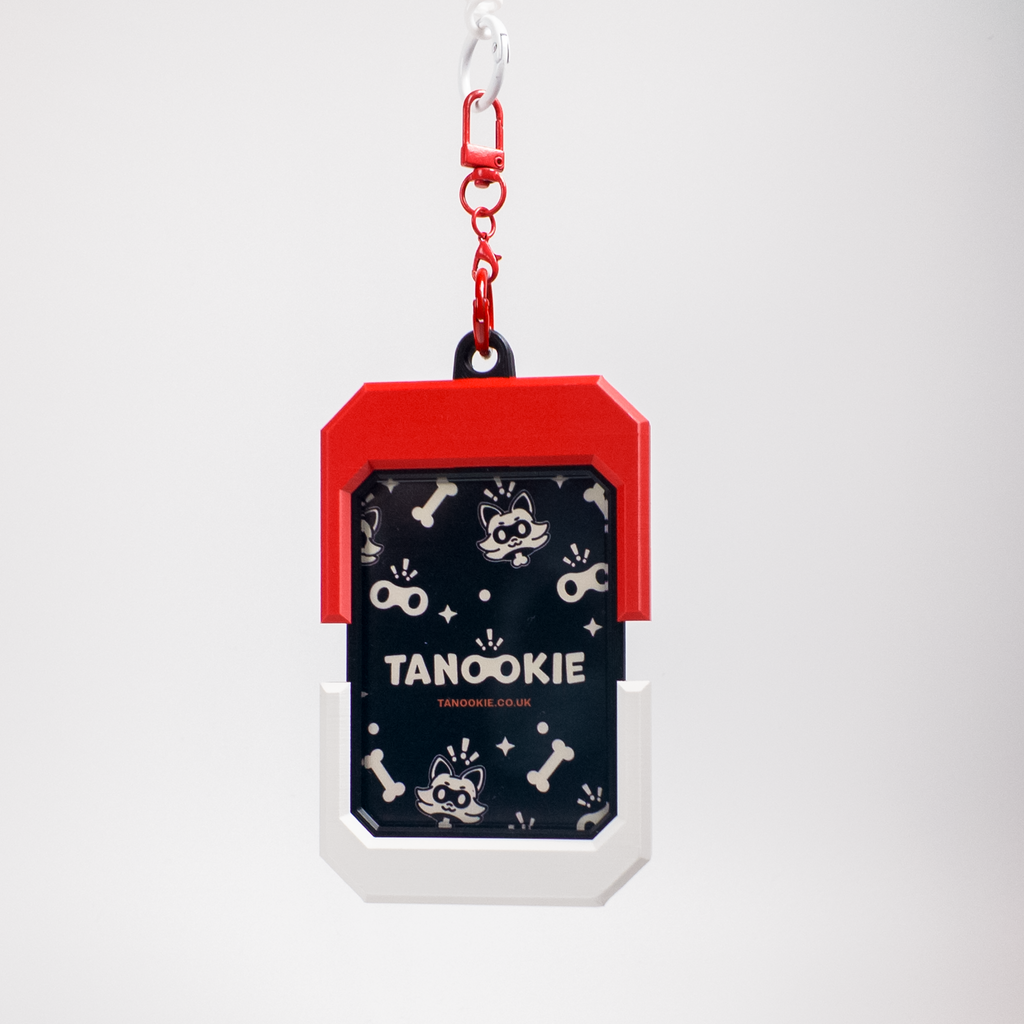 A pet ID tag or keychain-style accessory with a red top section, white bottom section, and a dark navy center panel printed with small white cartoon cat motifs and the text 'TANOOKIE' and 'STANLEY'. It hangs from a red carabiner on a light gray background.