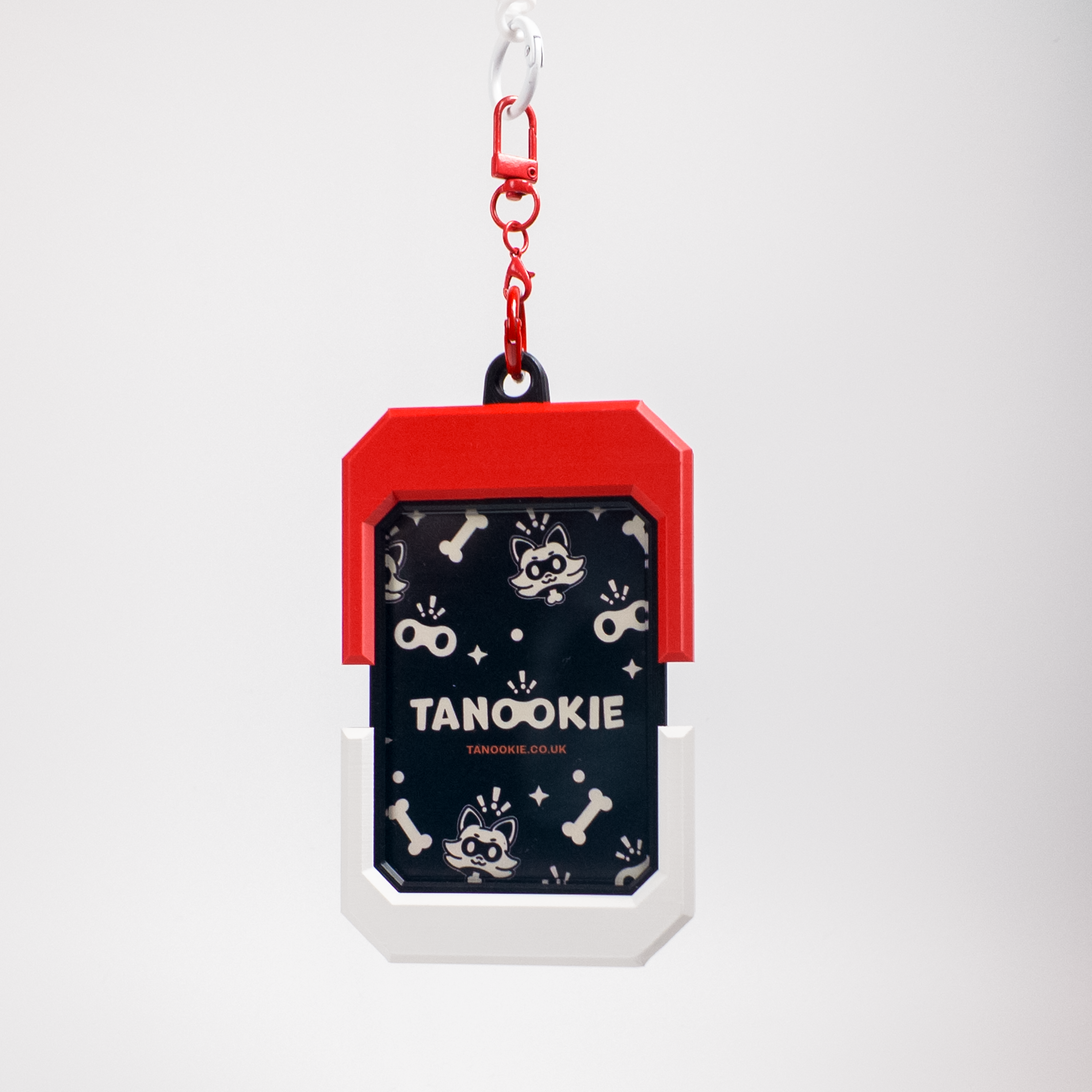 A pet ID tag or keychain-style accessory with a red top section, white bottom section, and a dark navy center panel printed with small white cartoon cat motifs and the text 'TANOOKIE' and 'STANLEY'. It hangs from a red carabiner on a light gray background.