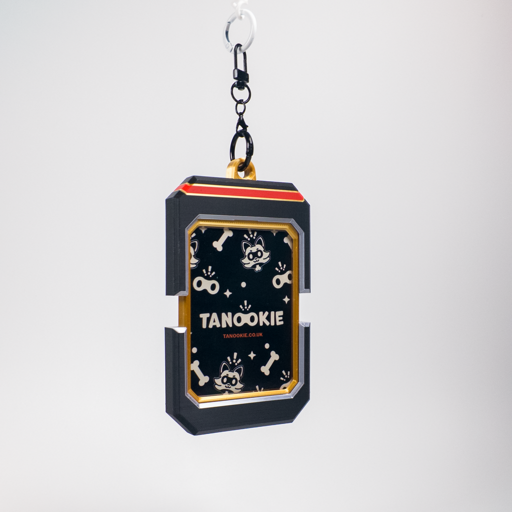 A black rectangular keychain or bag charm with a gold and red top trim, featuring a black front panel decorated with white cartoon dog illustrations and the text 'TANOOKIE' and 'TANOOKIE CLUB'. It hangs from a black chain and silver ring against a light gray background.