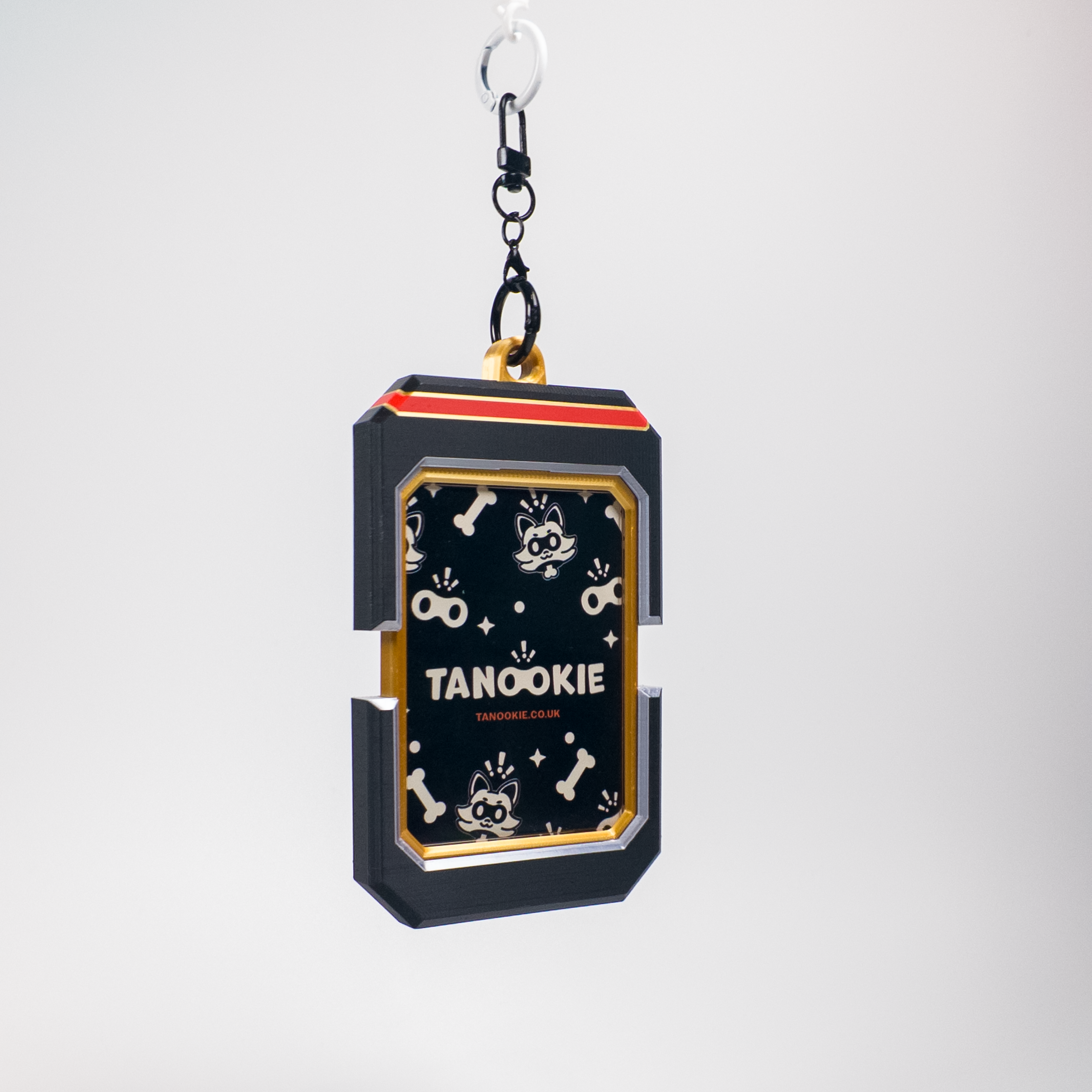 A black rectangular keychain or bag charm with a gold and red top trim, featuring a black front panel decorated with white cartoon dog illustrations and the text 'TANOOKIE' and 'TANOOKIE CLUB'. It hangs from a black chain and silver ring against a light gray background.